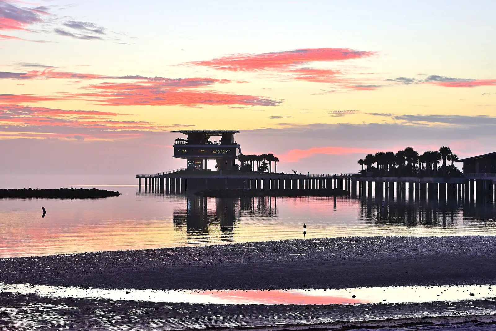 The St. Pete Pier silhouetted against a pink sunrise over Tampa Bay, seen from the shore at low tide.