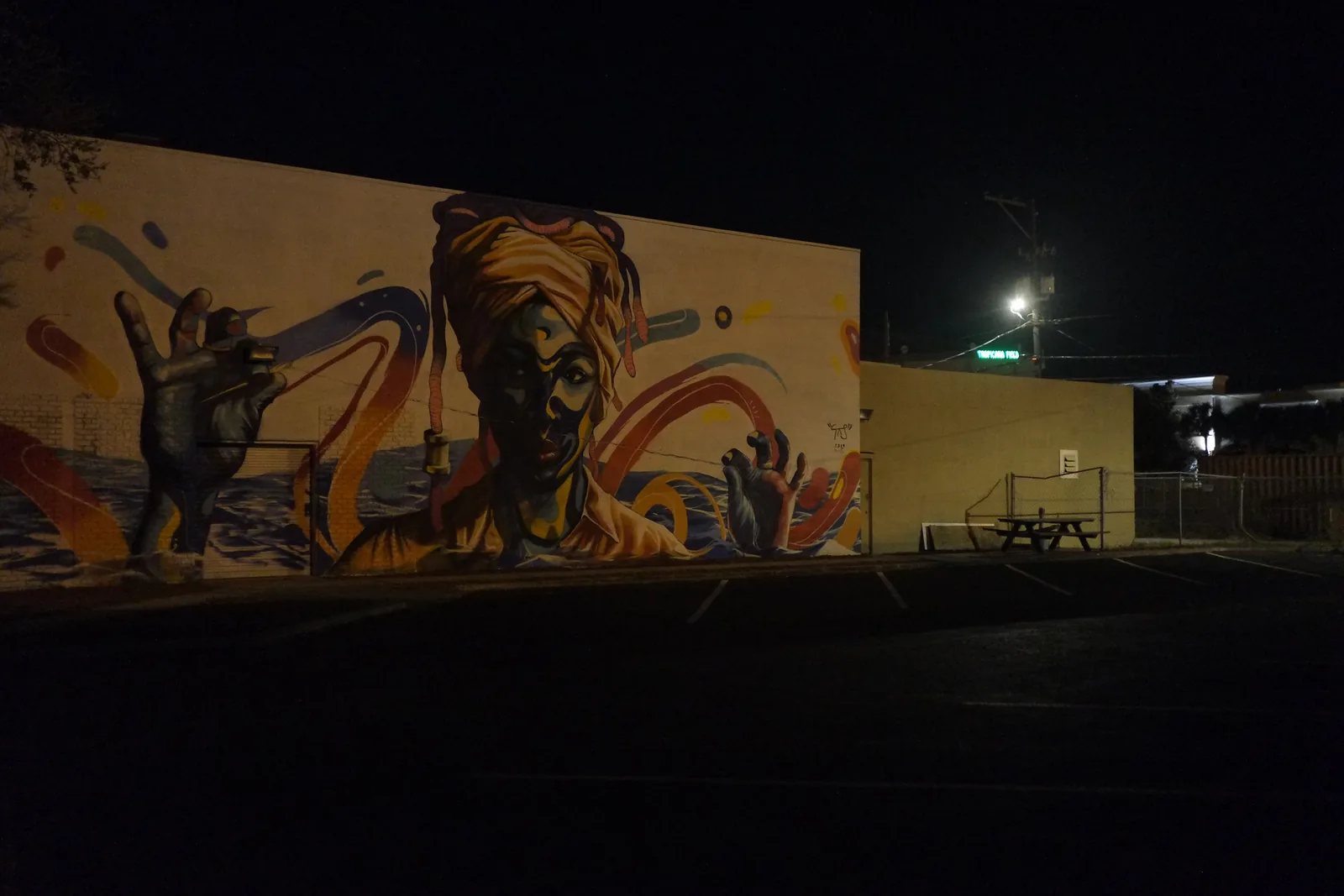 A large mural of a woman in a head wrap, lit at night on a building wall beside a parking lot in St. Petersburg.