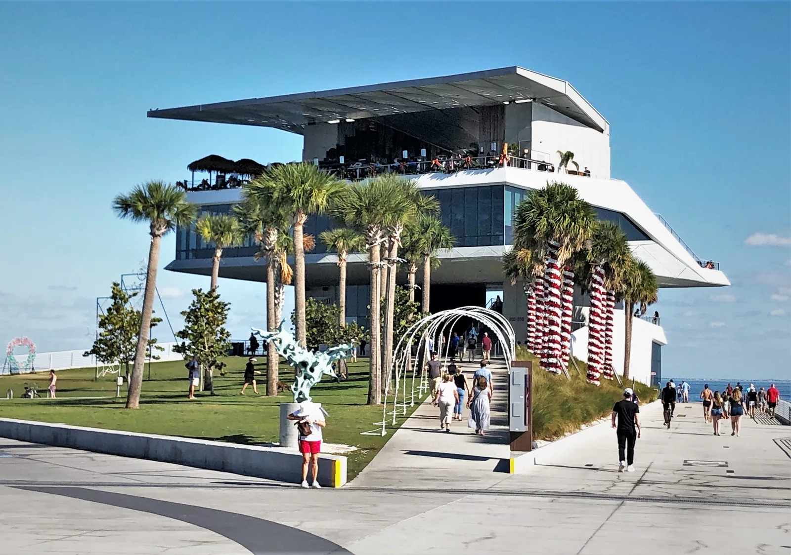 Visitors walk up to Pier Point, the building at the end of the St. Pete Pier, past palms and lawn on Tampa Bay.